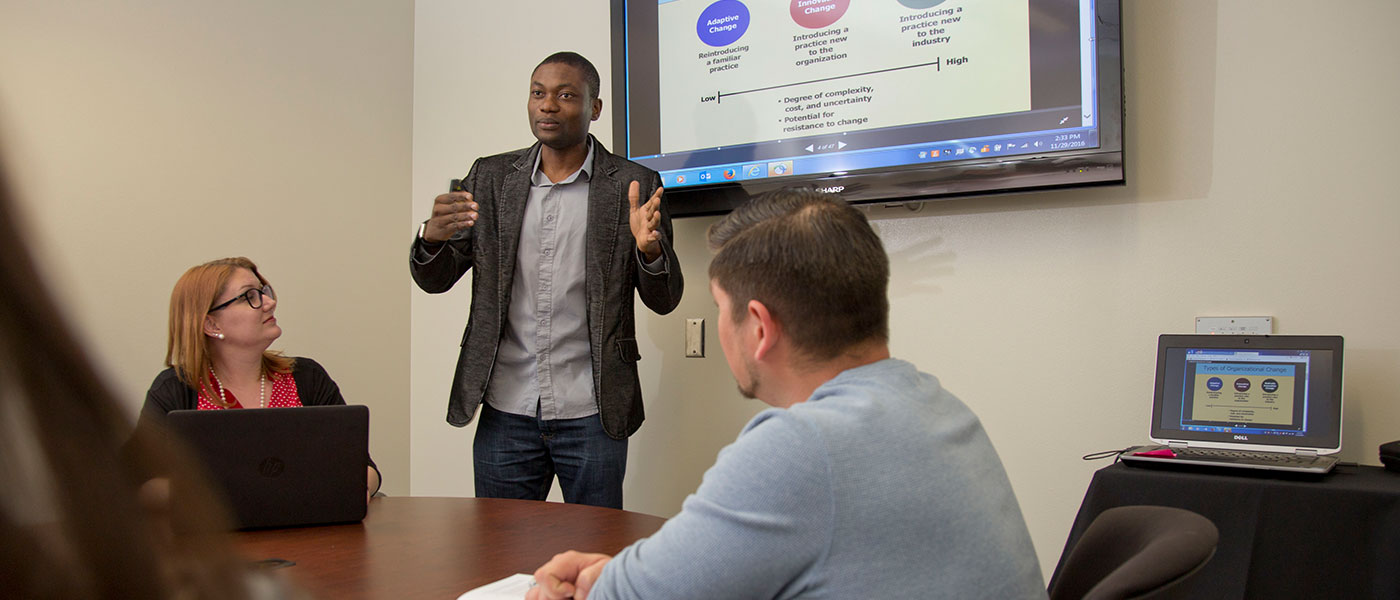 A man presenting to colleagues in a conference room in front of a monitor.