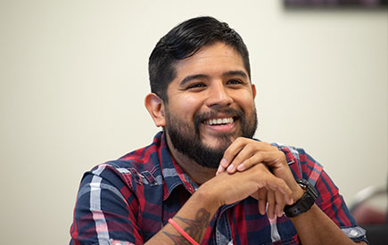 A man in the classroom smiling at his desk with his hands crossed.