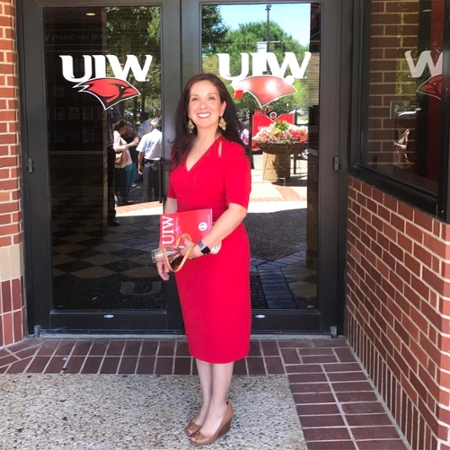 Jessica Davila-Burnett in a red dress standing outside of doors with the UIW logo
