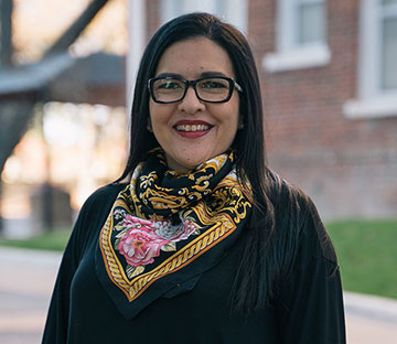 A smiling lady in glasses and a scarf standing outside.